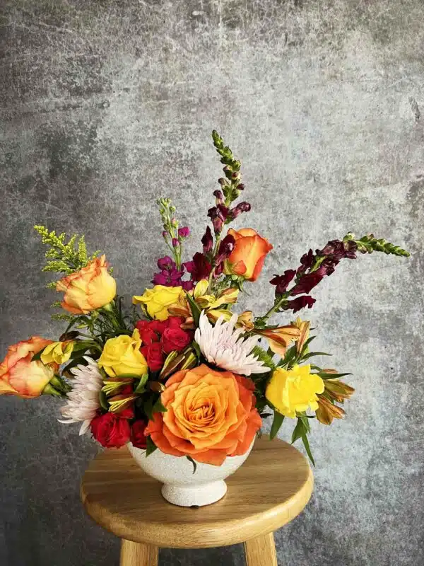 A vibrant "Sunsets" flower arrangement featuring orange Garden Roses, yellow Buttercups, Snapdragons, and other colorful blooms in a white vase, available in Washington, NC.
